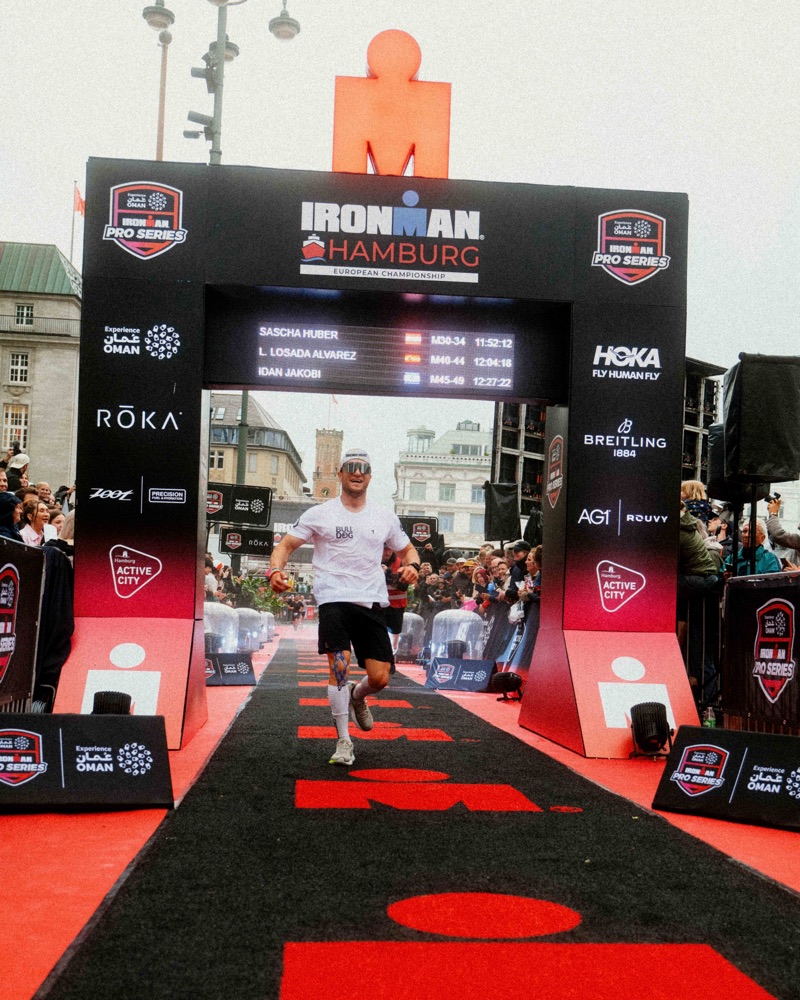 Sascha Huber läuft durch das Zielportal des IRONMAN Hamburg, die Anzeigetafel zeigt seinen Namen und die Zeit 11:52:12